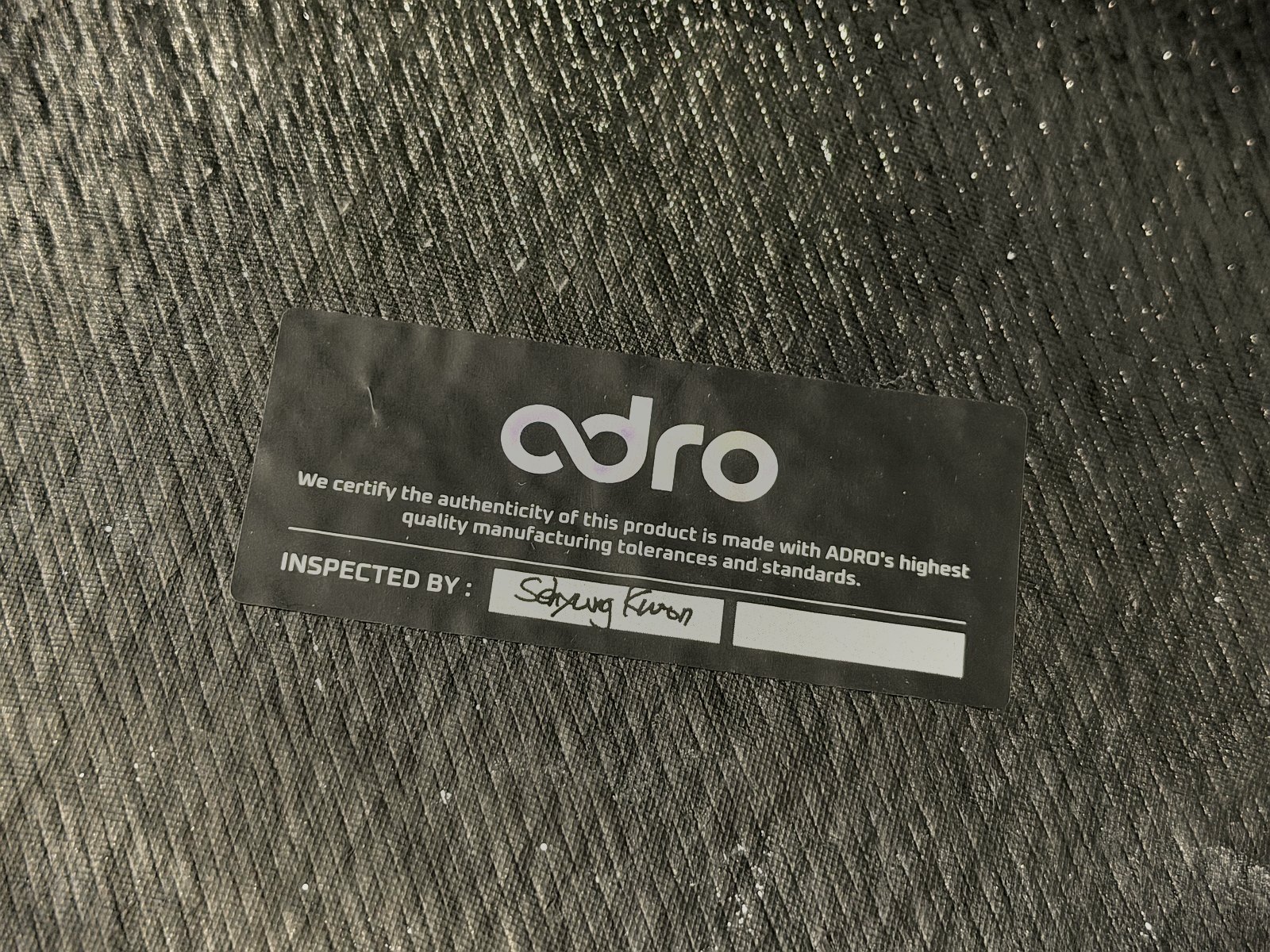 สติคเกอร์รับรอง ADRO ของแท้ด้านหลังชิ้นงานคาร์บอน — โฮโลแกรมพร้อมลายเซ็นผู้ตรวจ INSPECTED BY